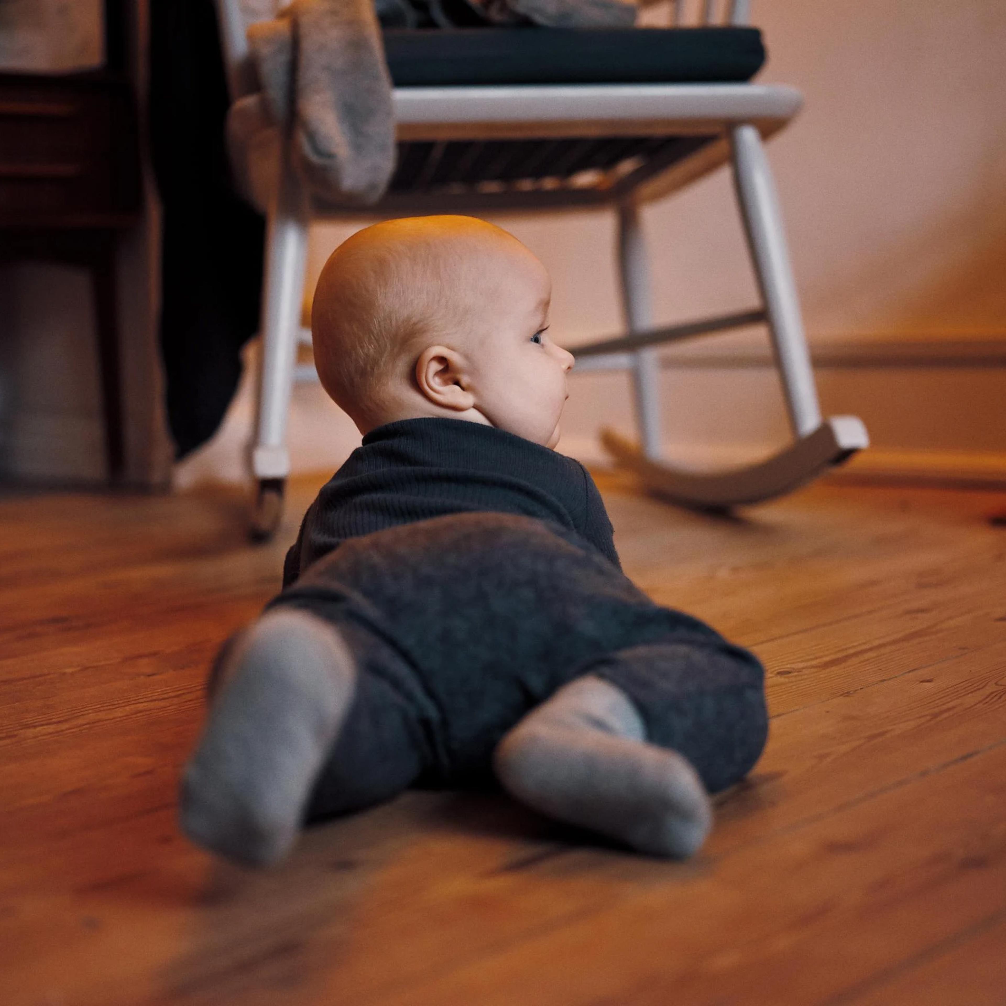 Et lille barn ligger på maven på et trægulv og kigger mod en hvid gyngestol. Barnet har mørkt tøj og sokker på, og billedet er taget i et hyggeligt rum med varme farver.