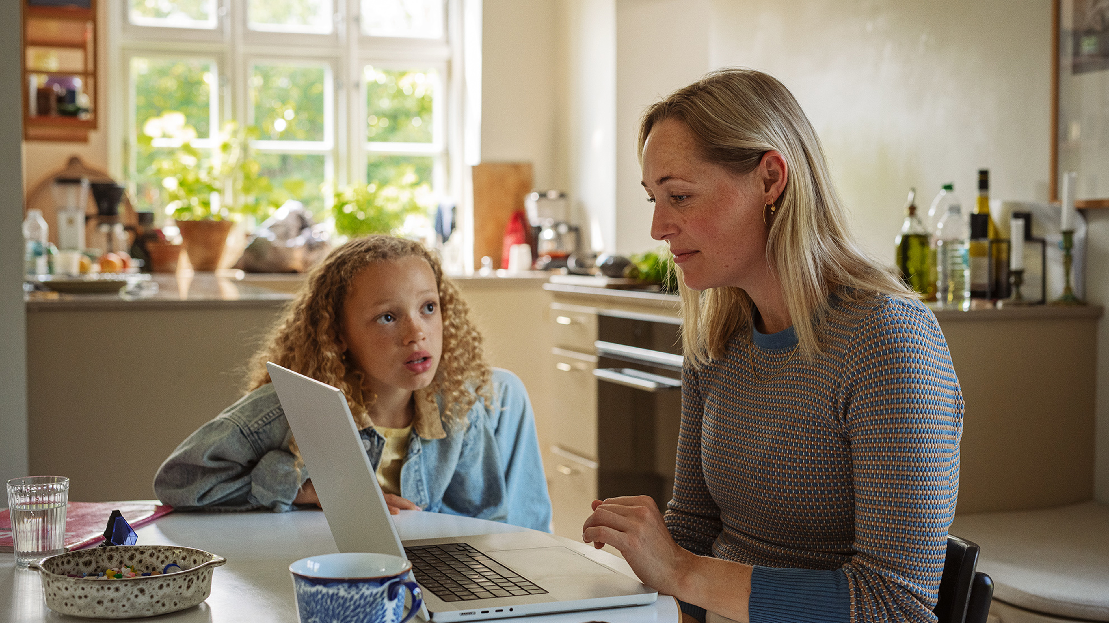 Mor arbejder på sin computer i køkkenet med sin datter ved siden af